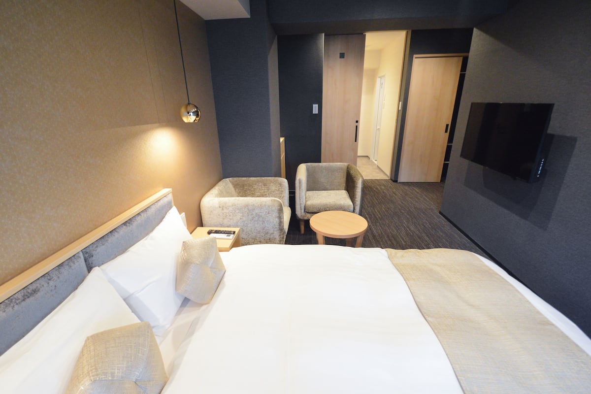 A hotel room with a bed, two armchairs, a small table, a TV, and a door leading to another room.