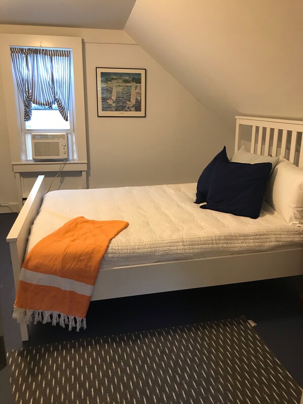 A bedroom with a bed, a window with curtains, an air conditioner, a framed picture on the wall, and a patterned rug. - Come enjoy Orleans and Chatham (Orleans)