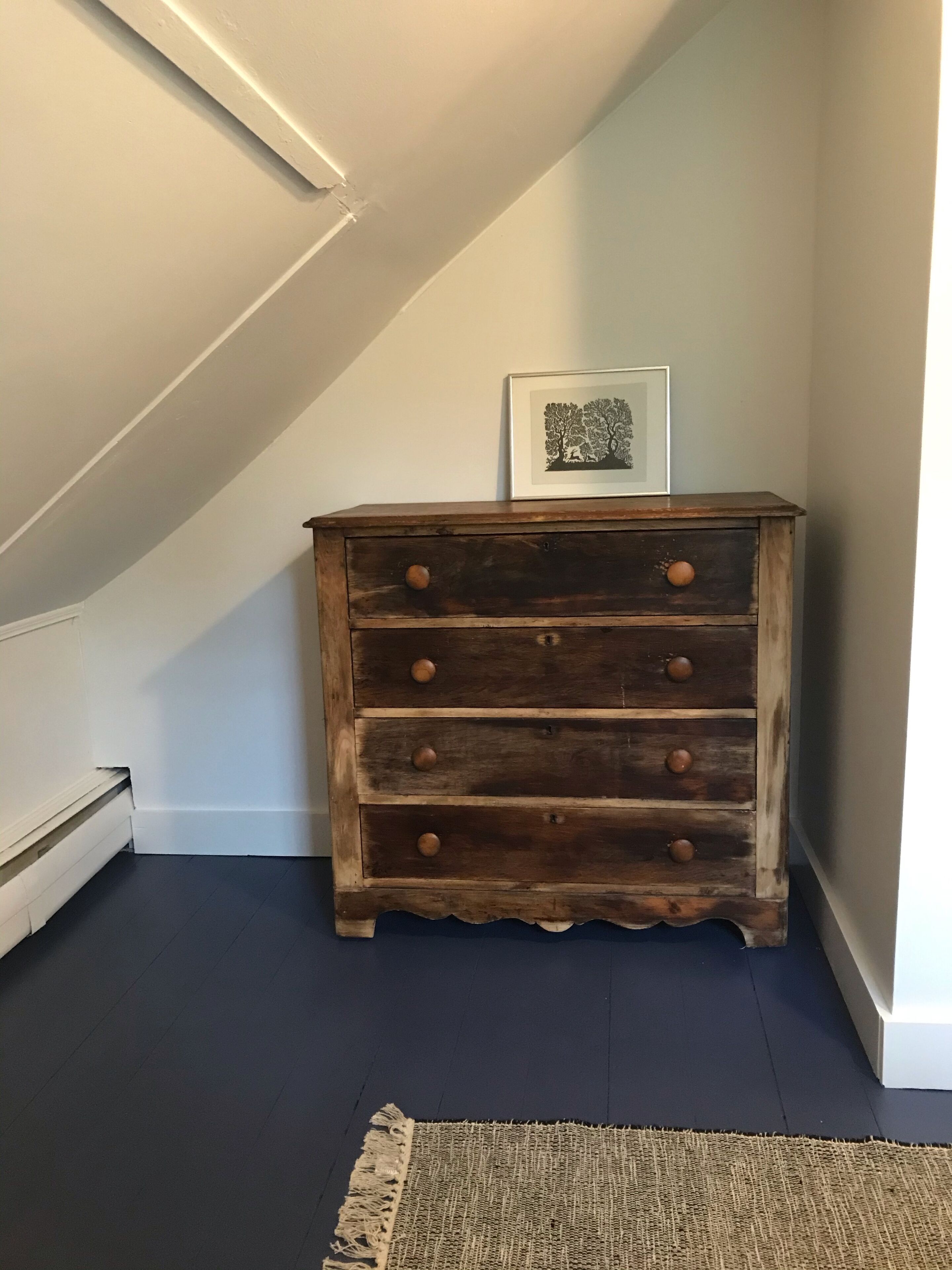 Une commode en bois avec des tiroirs et des boutons, un tableau encadré au-dessus, et un plancher foncé.
