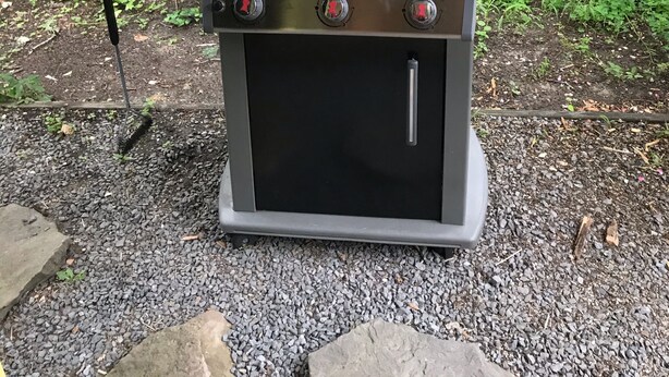 A Weber SPIFIRE grill with two side burners and a closed lid, set on a gravel surface with a metal table in front, surrounded by greenery and flowers.