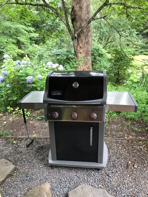 Ein Weber SPIFIRE Grill mit zwei Seitenbrennern und geschlossenem Deckel, auf einer Schotterfläche mit einem Metalltisch davor, umgeben von Grünpflanzen und Blumen.