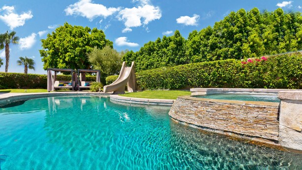 A clear swimming pool with a slide, surrounded by lush greenery and a stone wall. - Home Theater, Arcade games, Water Slide, Lagoon-Style Pool & Extra-Large Spa (Indio)