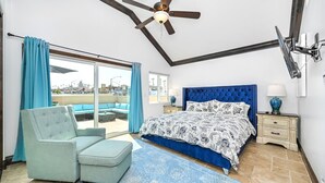 Een slaapkamer met een groot bed, een blauwe fauteuil, een plafondventilator en uitzicht door een glazen deur.