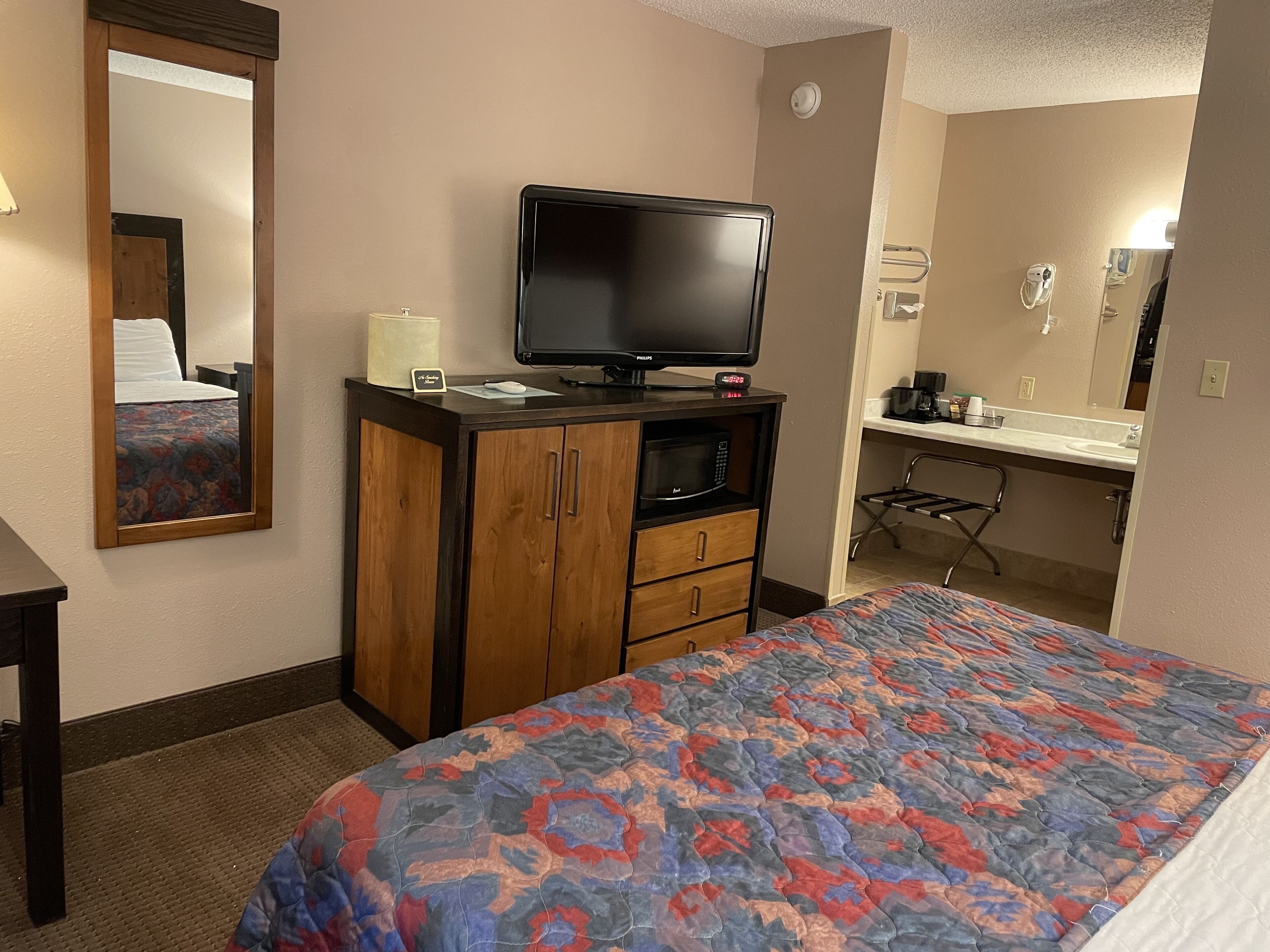 A hotel room with a bed, a television on a wooden cabinet, a microwave, a small table with a lamp, and a bathroom with a sink and mirror.