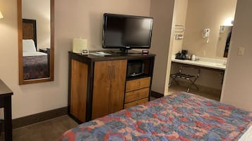 A hotel room with a bed, a television on a wooden cabinet, a microwave, a small table with a lamp, and a bathroom with a sink and mirror.