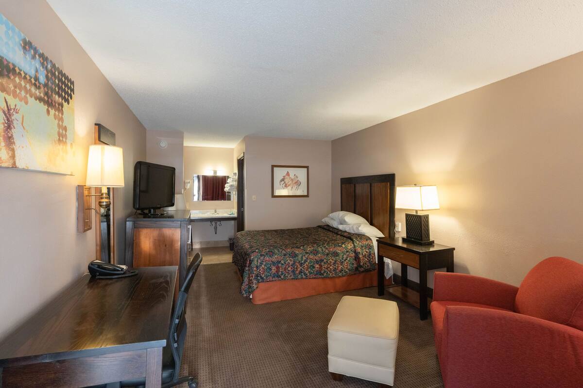 Una habitación de hotel con cama, escritorio con teléfono, una silla, televisor y lámpara.