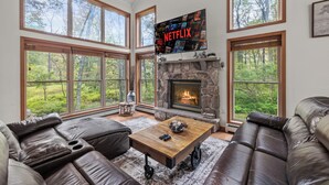 Una sala de estar con chimenea de piedra, ventanas amplias, sofás de cuero y una pantalla de Netflix.