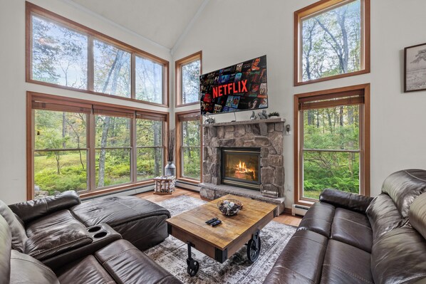 Una sala de estar con chimenea de piedra, ventanas amplias, sofás de cuero y una pantalla de Netflix.
