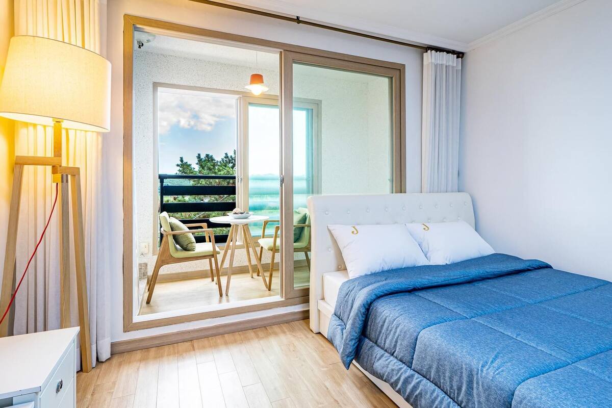 A bedroom with a bed, a nightstand, a lamp, and a view of a balcony with a table and chairs.