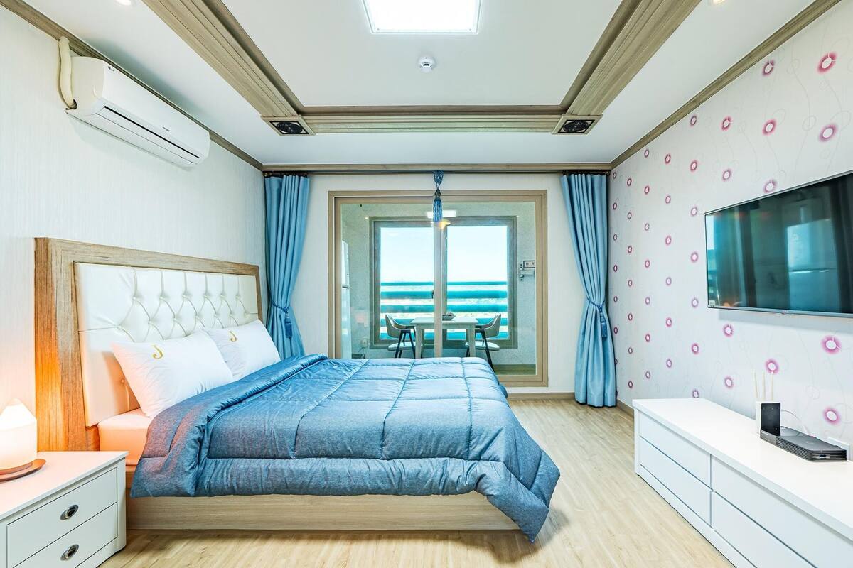 A bedroom with a bed, a TV, a desk, and a view of the beach.