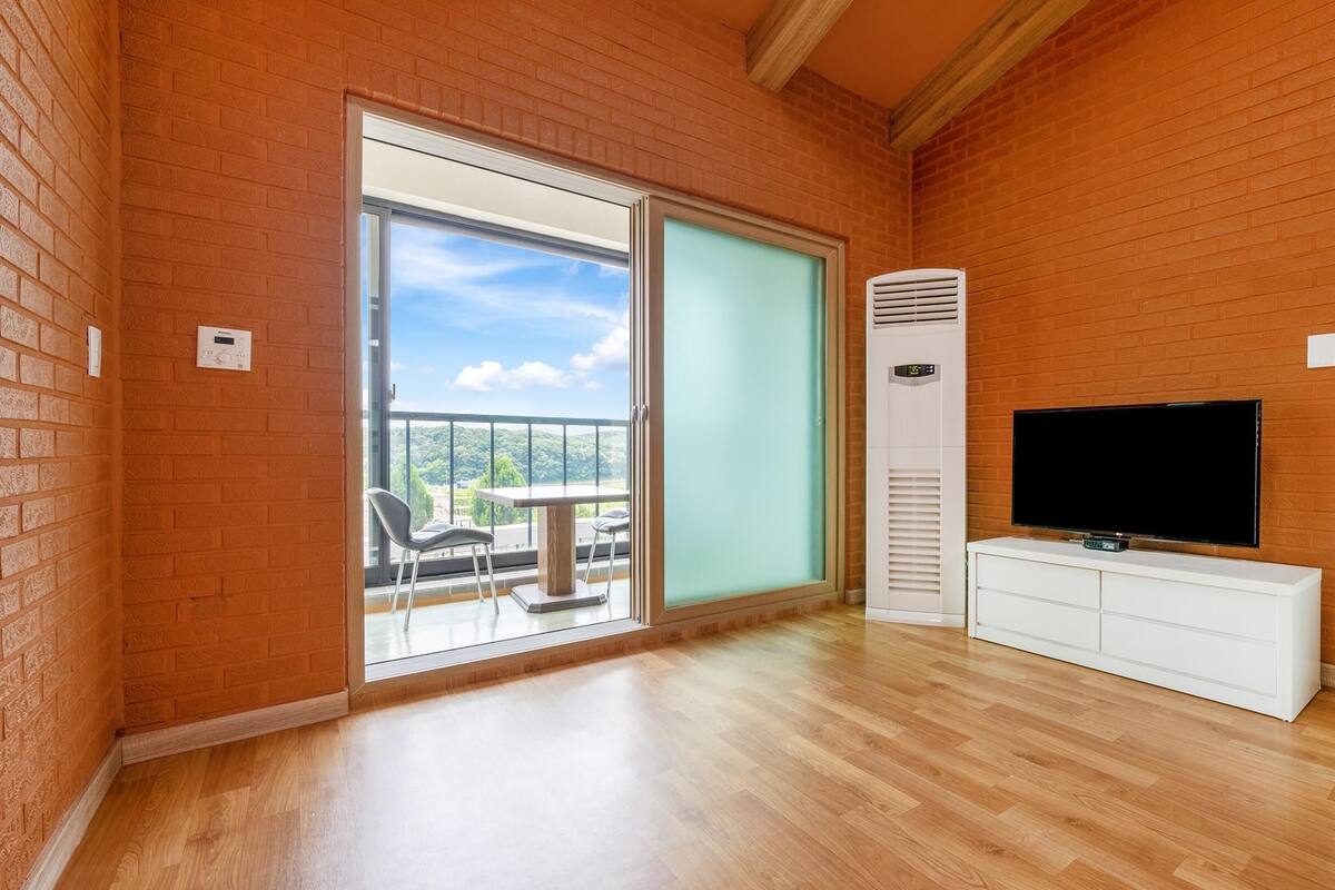 A room with a wooden floor, a TV on a white cabinet, a sliding glass door leading to a balcony, and a brick wall.