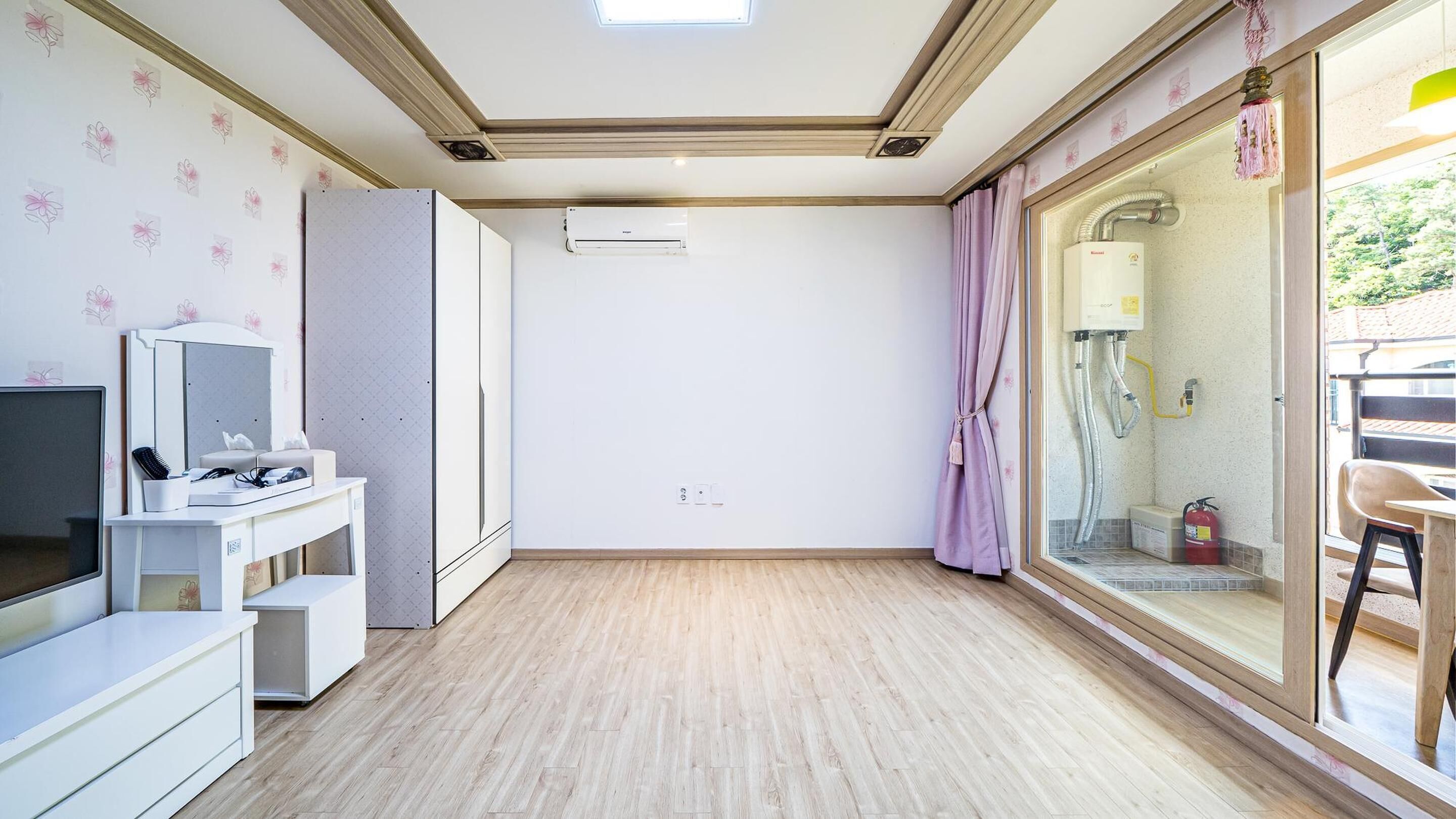 A room with a wooden floor, a white desk with a TV, a wardrobe, and a bathroom with a shower and toilet visible through a glass door.