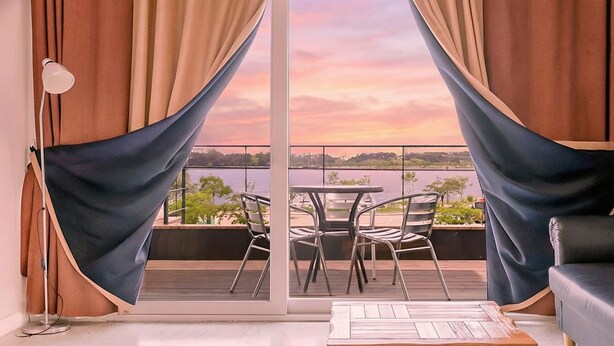 A balcony with a table and chairs, curtains, and a view of a body of water during sunset.