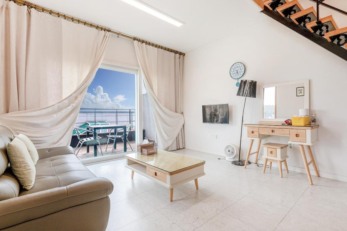 Ein modernes Wohnzimmer mit Sofa, Couchtisch, Schreibtisch mit Stuhl, Ventilator und einer Treppe.