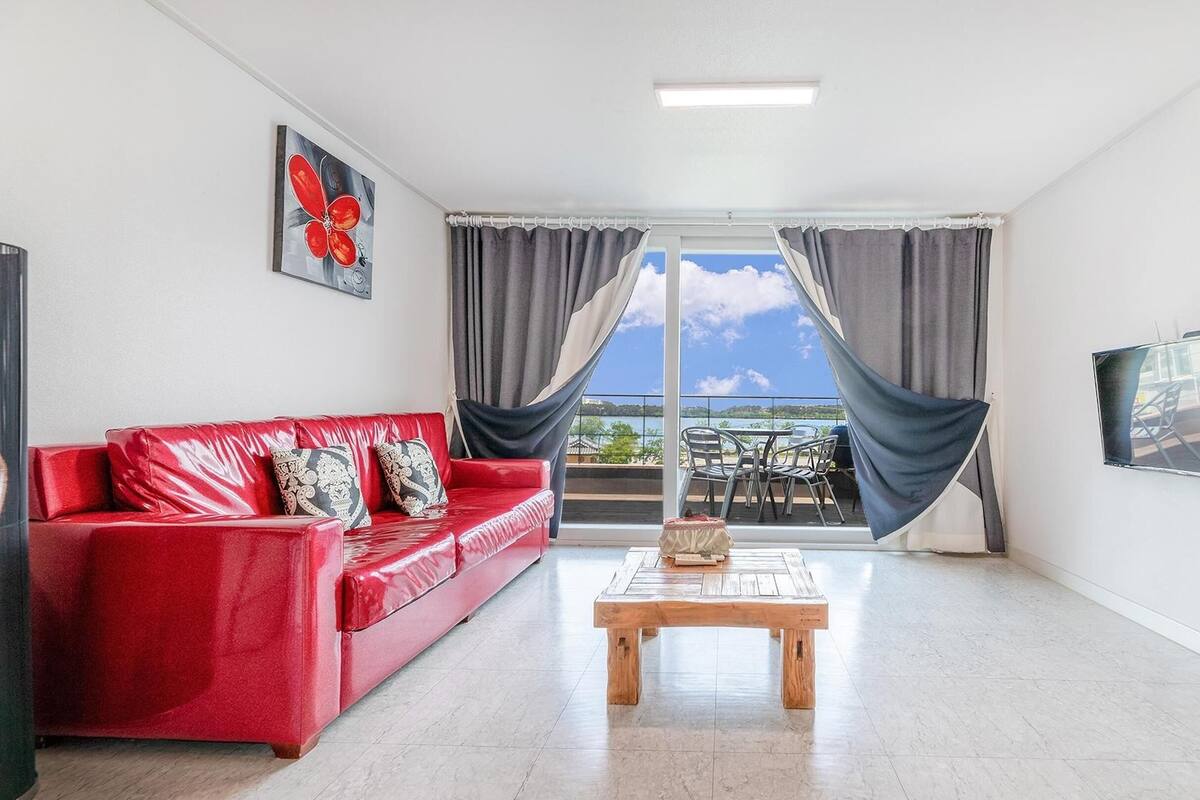 Ein Zimmer mit einem roten Ledersofa, einem Holztischchen, einem Fernseher an der Wand und Blick ins Freie durch große Fenster.