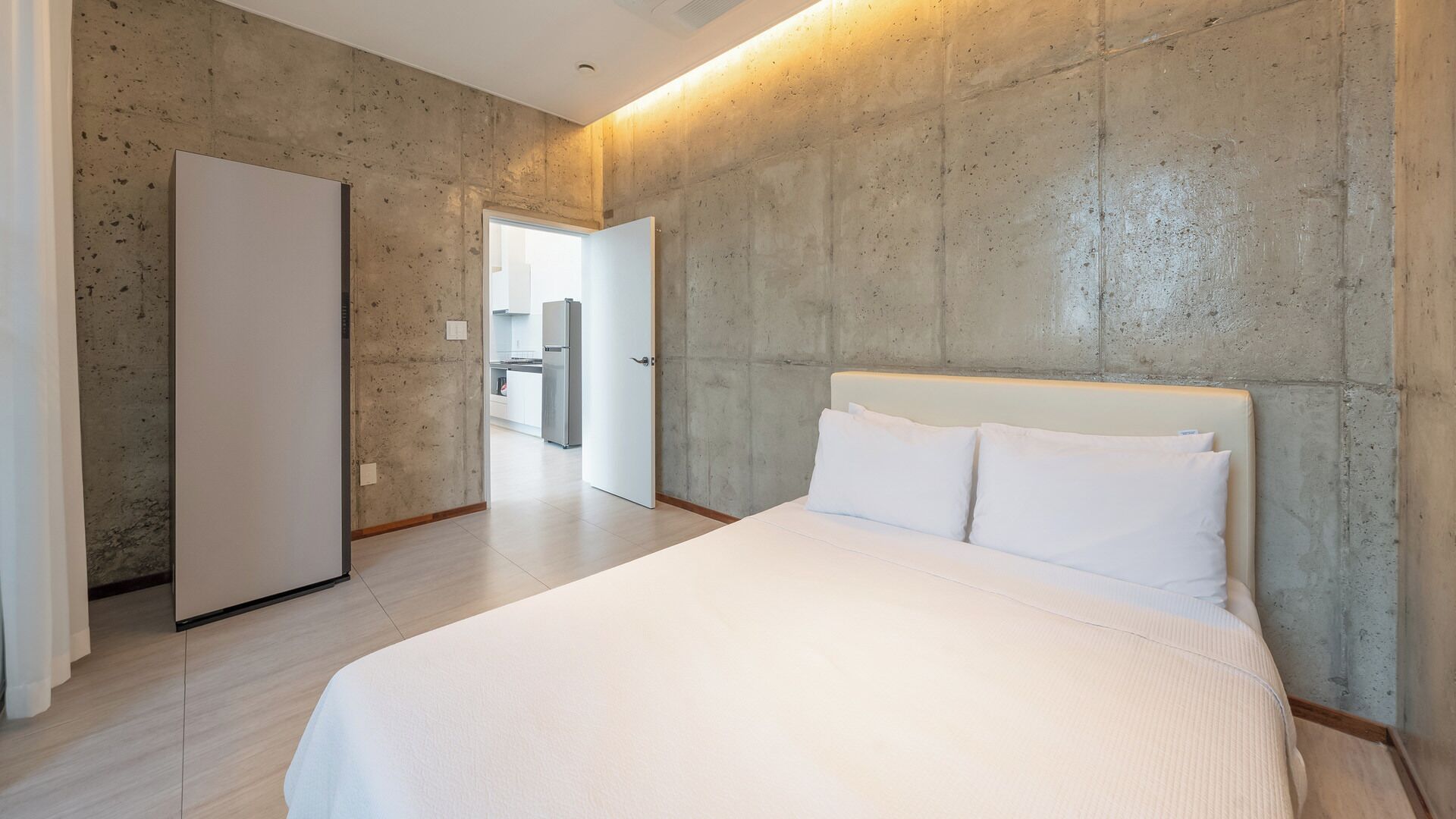 A bedroom with a large bed, a wardrobe, and a kitchen area visible through an open door.