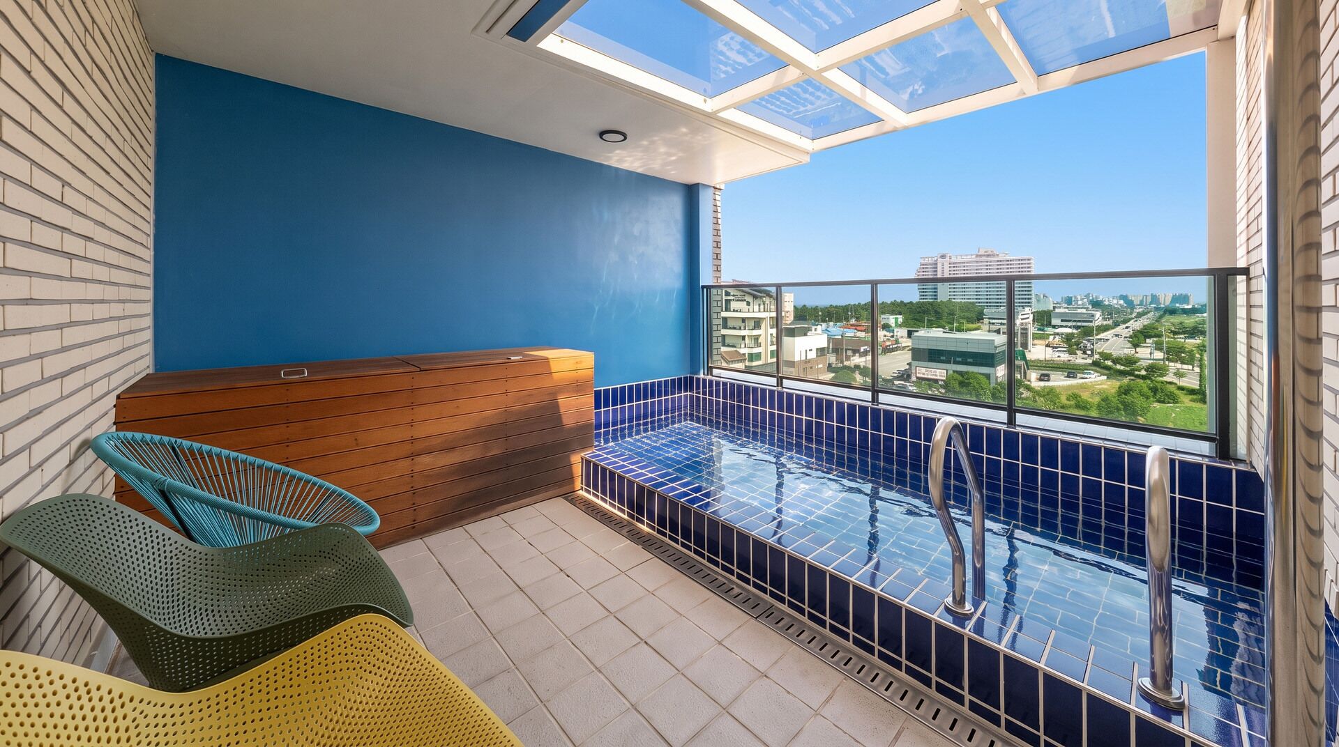 A rooftop terrace with a blue-tiled pool, wooden seating, and a view of the cityscape.