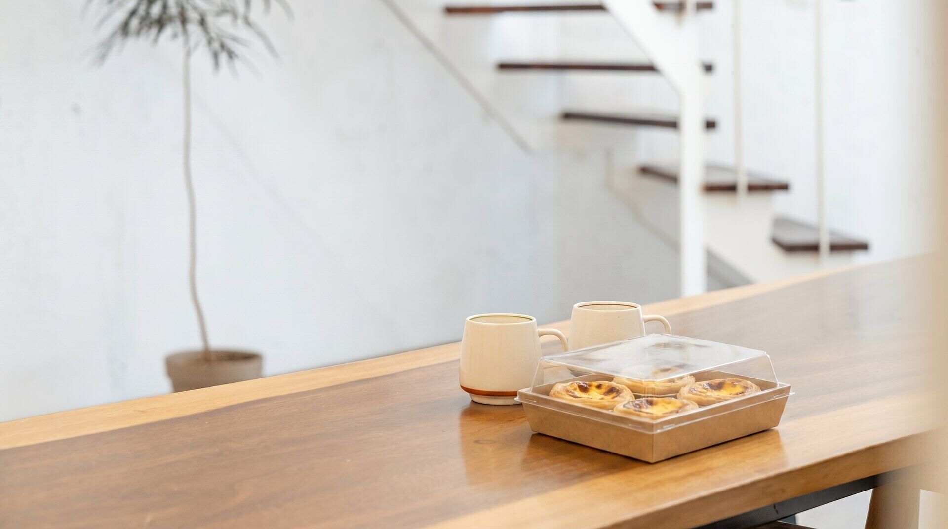 A wooden table with a box of Portuguese custard tarts and two ceramic mugs.