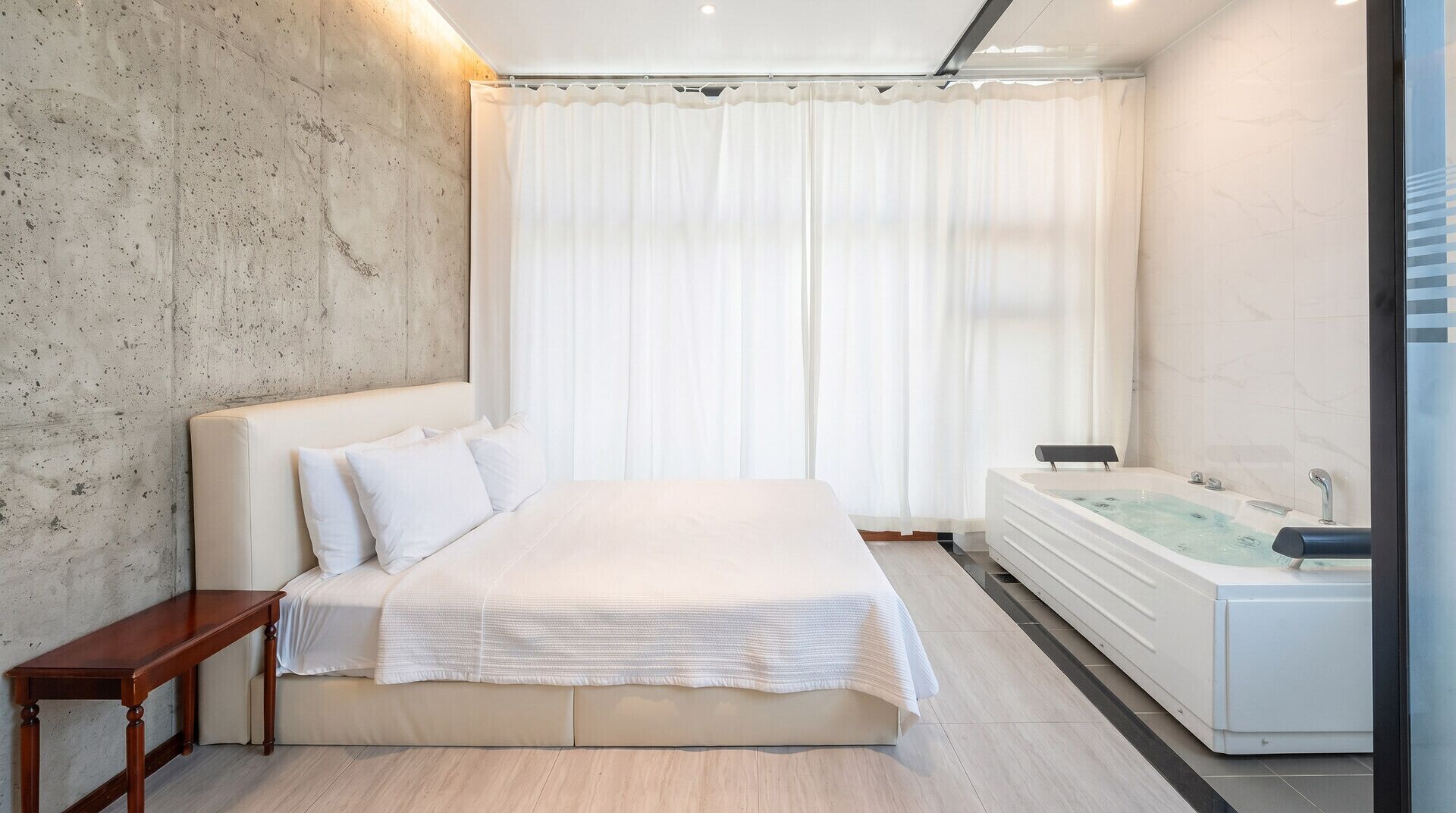 Ein modernes Schlafzimmer mit einem großen Bett, einem Holz-Nachttisch, einem weißen Bad mit einer Badewanne und einer Betonwand.