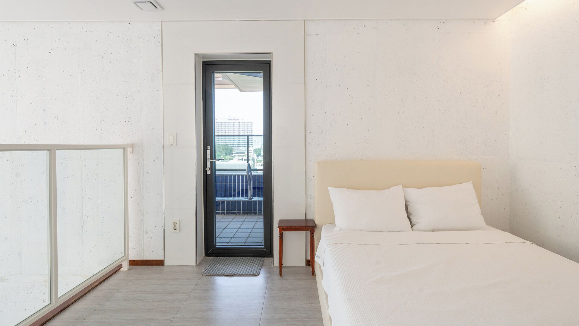 A bedroom with a bed, a nightstand, a door leading to a balcony, and a view of buildings outside.