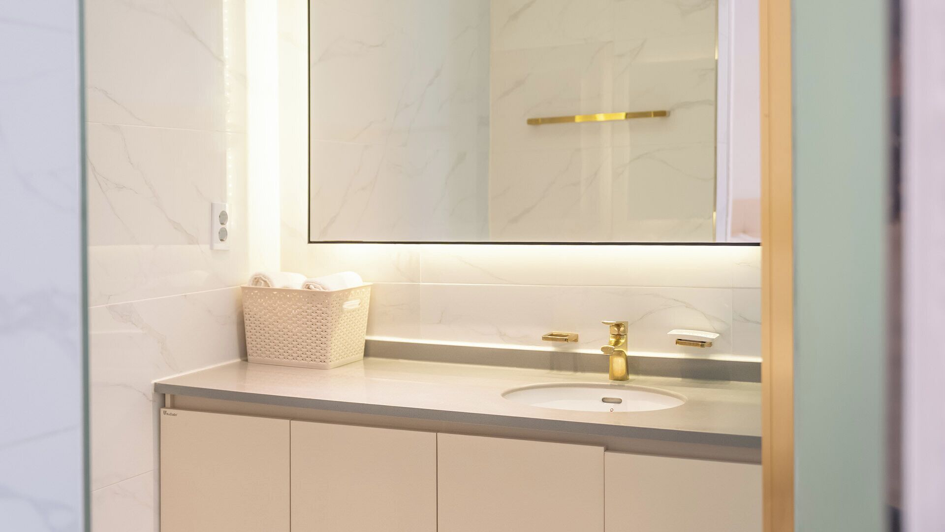 Une salle de bain avec un mur recouvert de carreaux de marbre, un lavabo blanc, un robinet doré et un miroir au-dessus du lavabo.