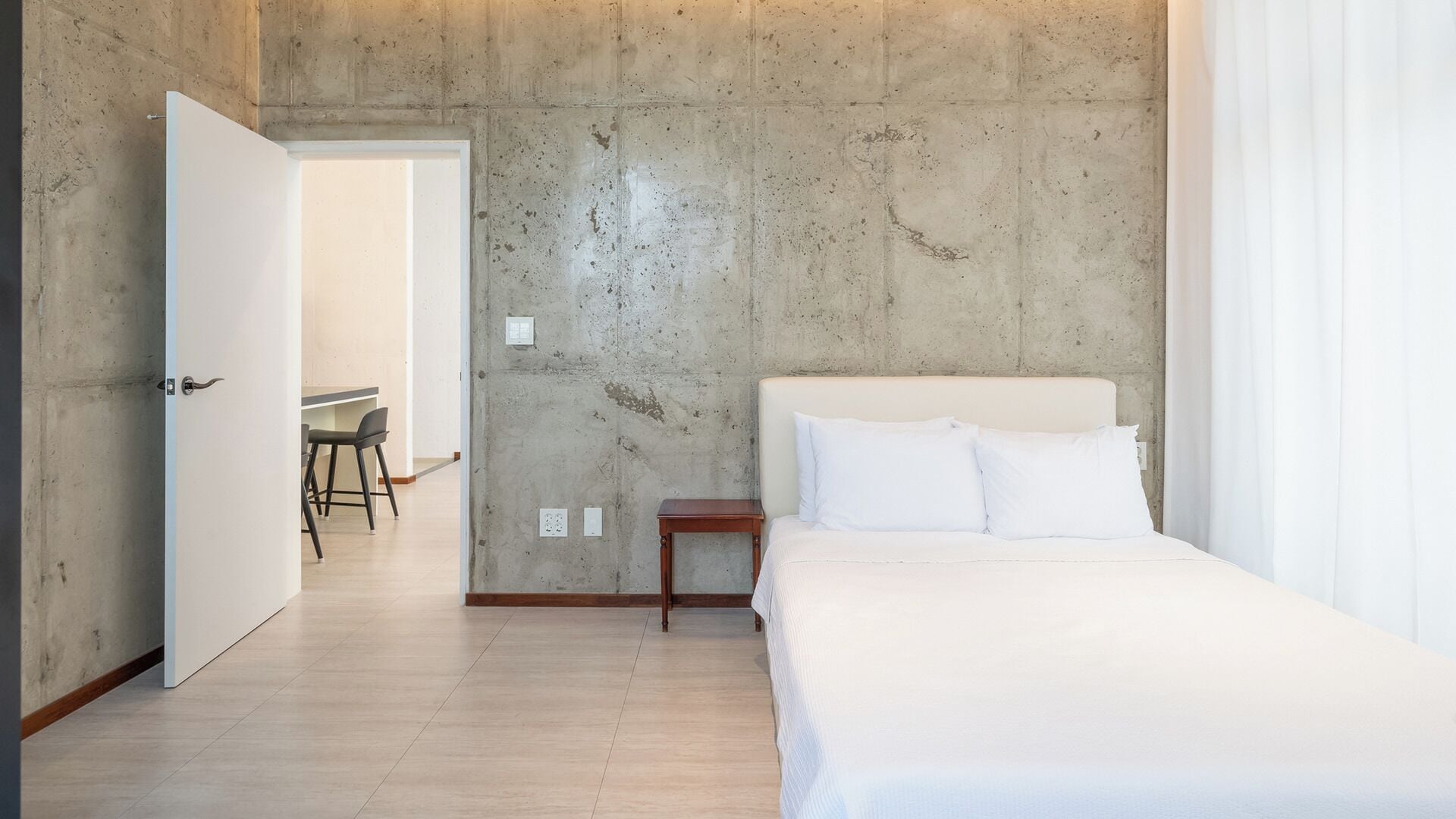 A bedroom with a bed, a nightstand, and a chair in a modern room with concrete walls and wooden flooring.