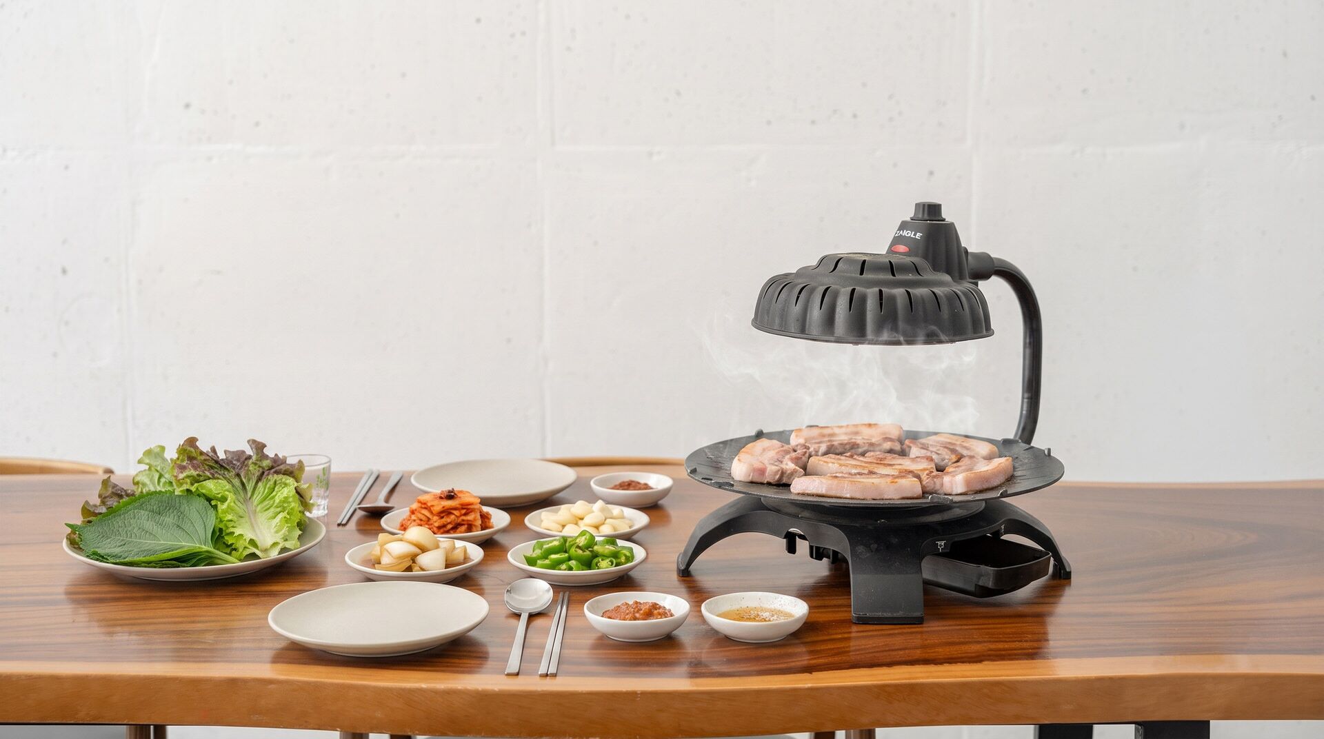 A table with a portable grill, various side dishes, and a white wall background.