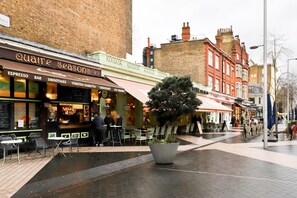 Une rue avec des terrasses, un café appelé « Quatre Saisons » et un grand bac à fleurs contenant un arbre.