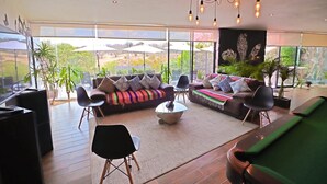 Amplia sala de estar con un sofá grande, una mesa de centro y una mesa de billar. Hay varias sillas y una mesa de comedor. La habitación tiene ventanas grandes con vistas al exterior.