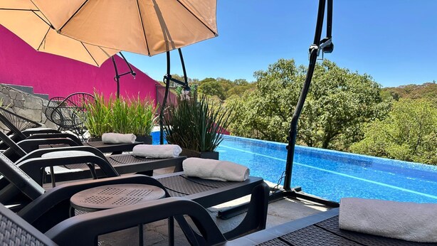 Zona de piscina con sillones y sombrillas, una pared de piedra y vistas a árboles y colinas.