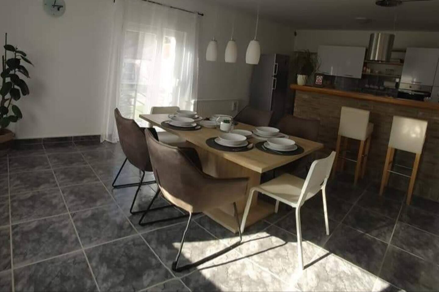 Een eethoek met een tafel gedekt voor vier personen, omringd door stoelen, in een kamer met een betegelde vloer en een keuken op de achtergrond.