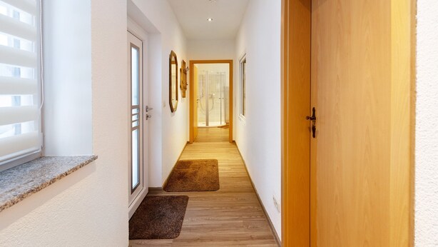 A hallway with wooden flooring, white walls, a wooden door, and a window with blinds.