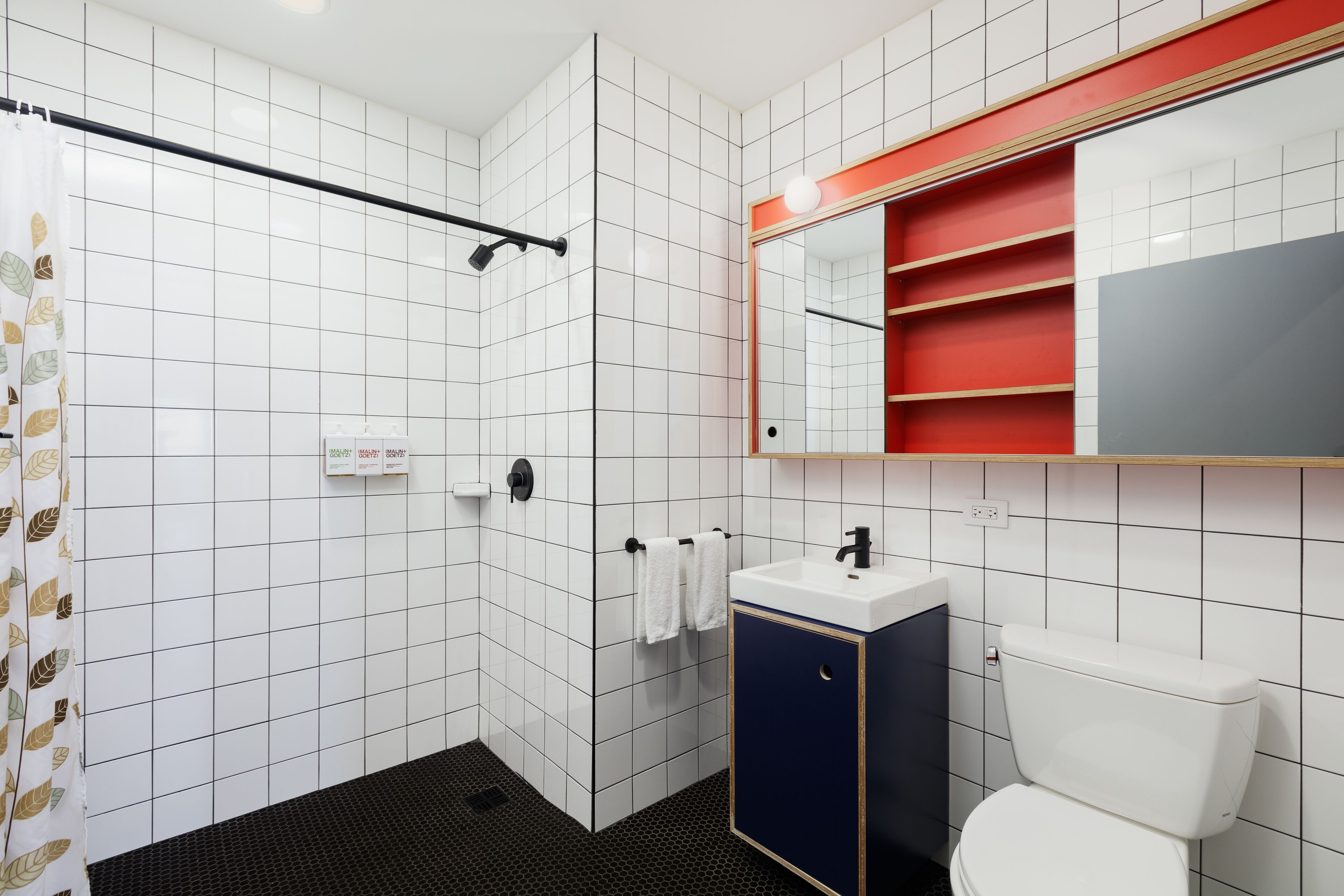 Un baño moderno con paredes de azulejos blancos, zona de ducha negra, inodoro blanco y lavamanos con gabinete azul.