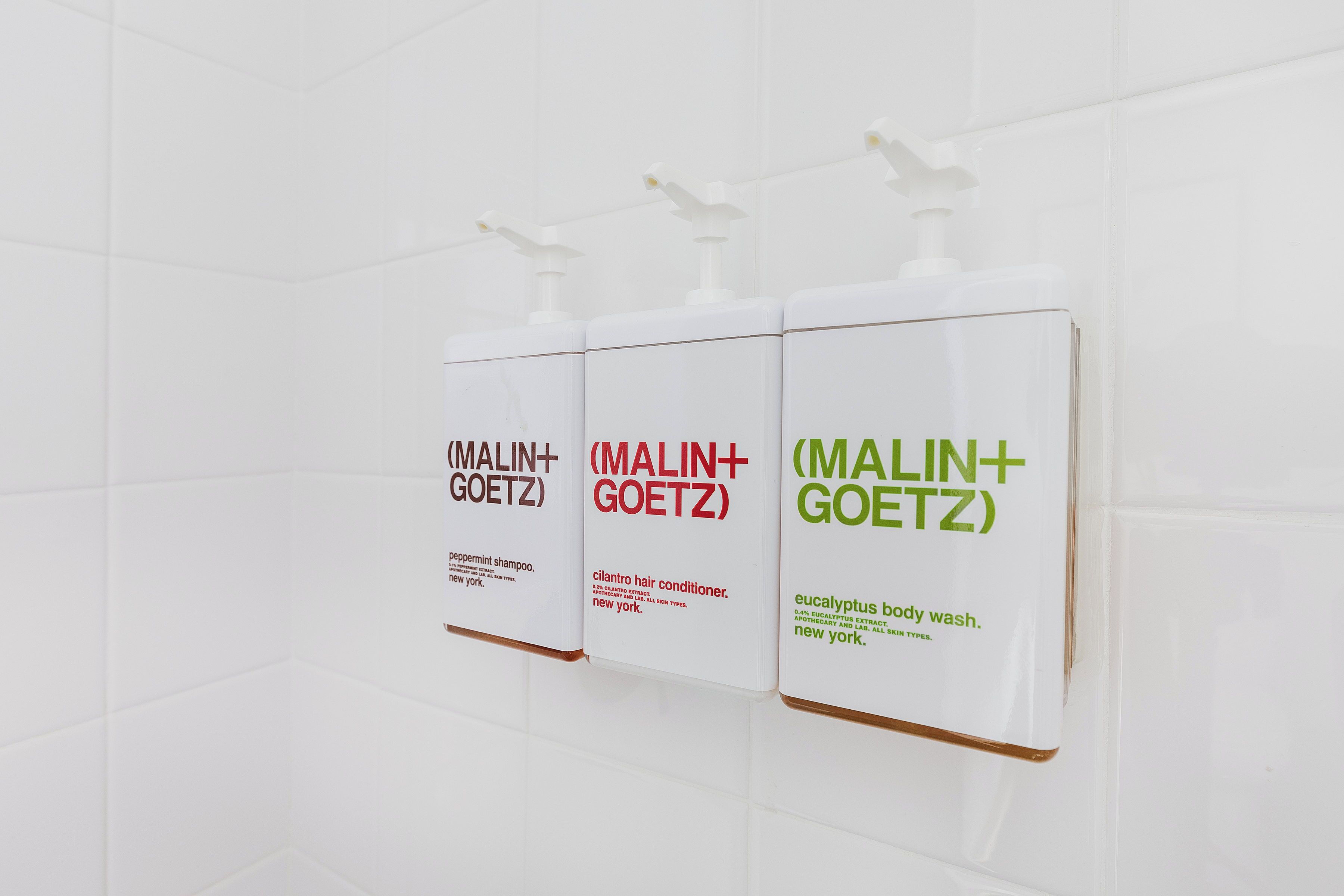 Drie witte dispensers met (MALIN+GOETZ) op een witte betegelde muur.
