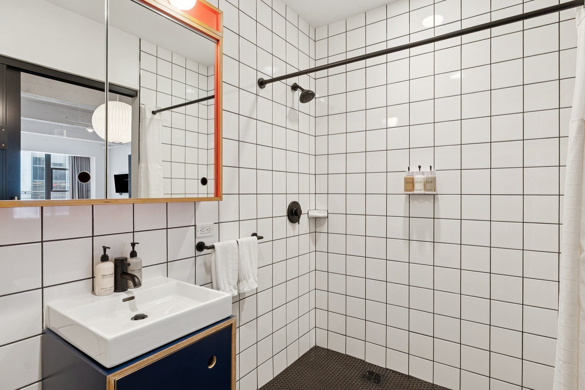 Un baño moderno con ducha de azulejos blancos, lavamanos con espejo y artículos de aseo.