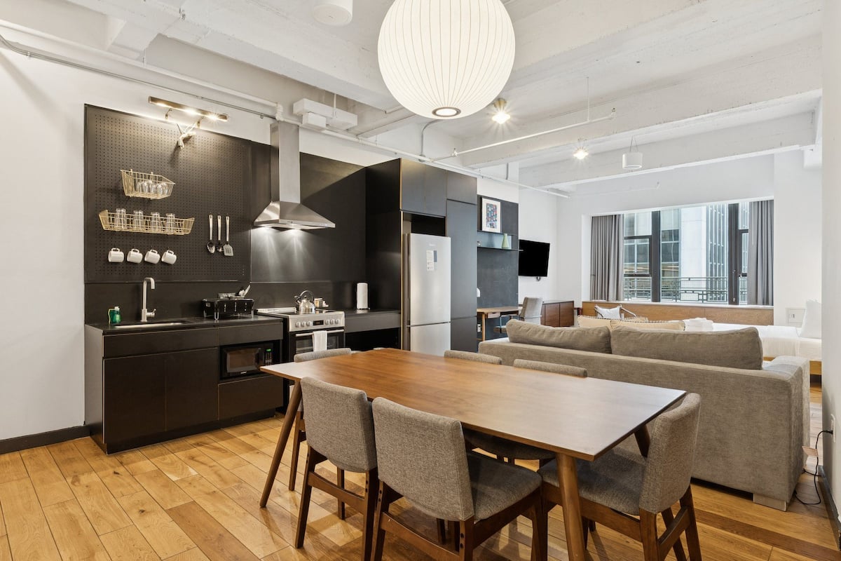A modern kitchen with a dining area, featuring a wooden table and chairs, a large window, and a dark backsplash.
