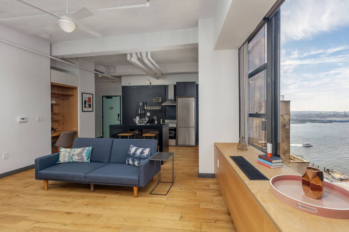 A modern apartment with a living area, kitchenette, and a large window overlooking a body of water.