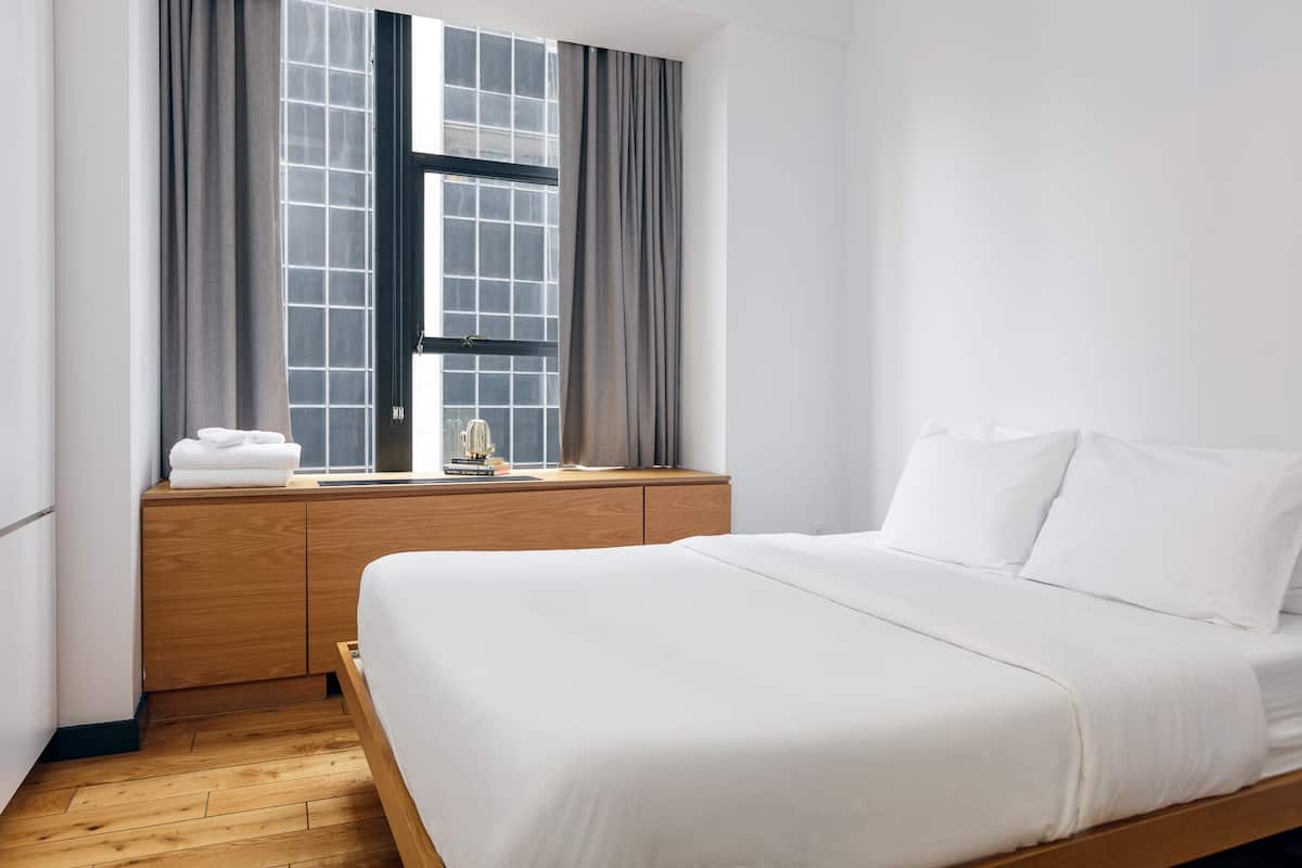 A minimalist hotel room with a wooden bed frame, a wooden nightstand, and a window with grey curtains.