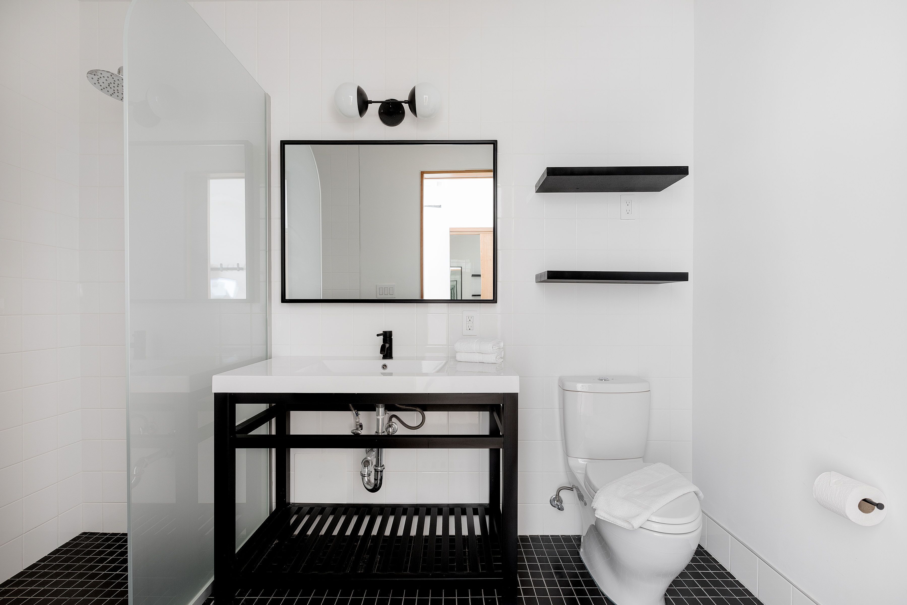 A modern bathroom with a white sink, black countertop, and a mirror above it. There is a toilet with a white lid and a roll of toilet paper on the wall.