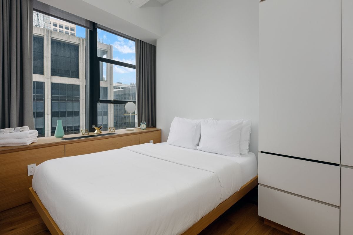Habitació d'hotel moderna amb un llit gran, una finestra amb cortines, una taula de nit amb una làmpada i un armari.
