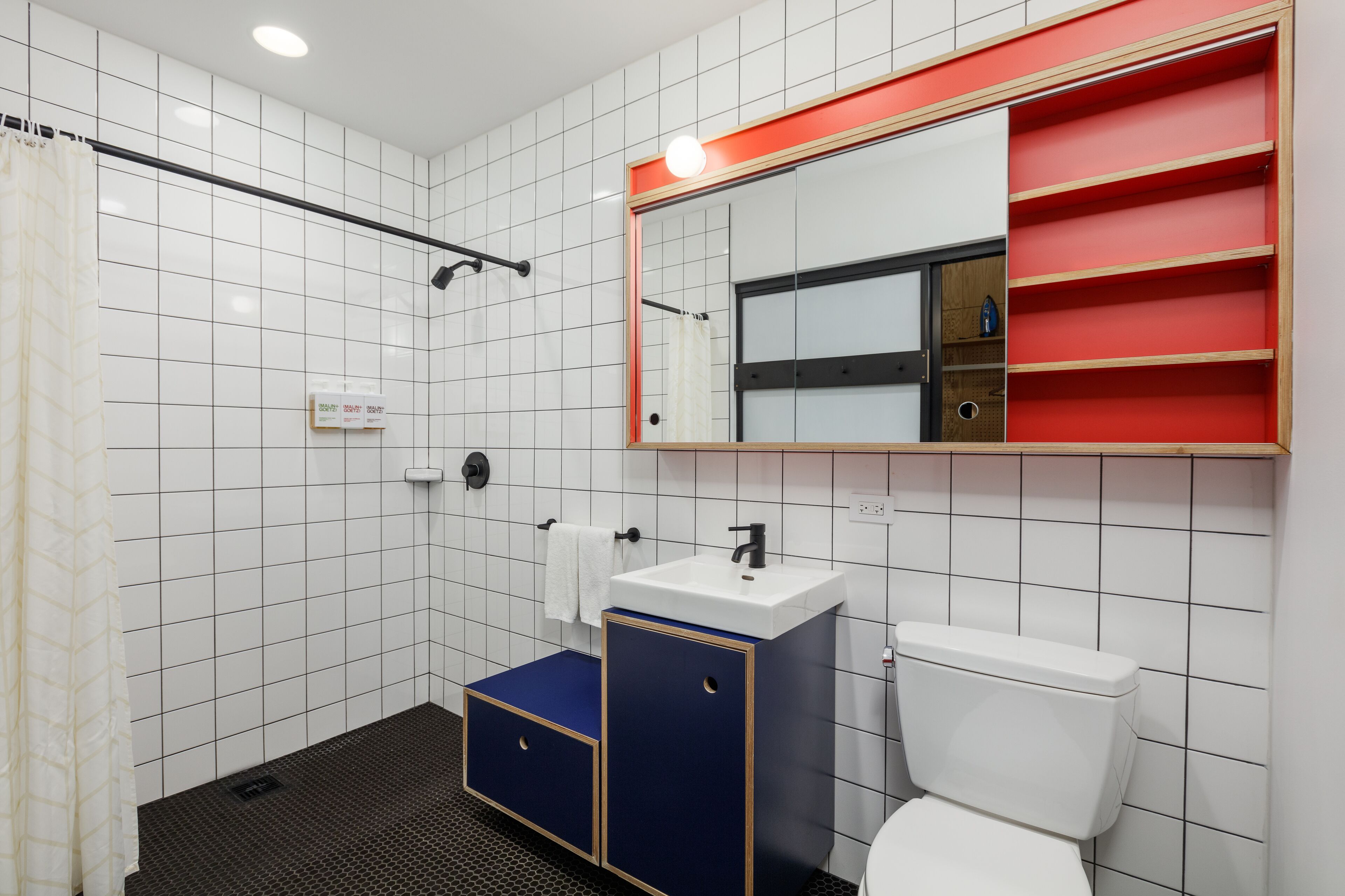 Un baño moderno con paredes de azulejos blancos, un inodoro blanco, un lavabo azul y negro, un espejo con marco rojo y una zona de ducha.