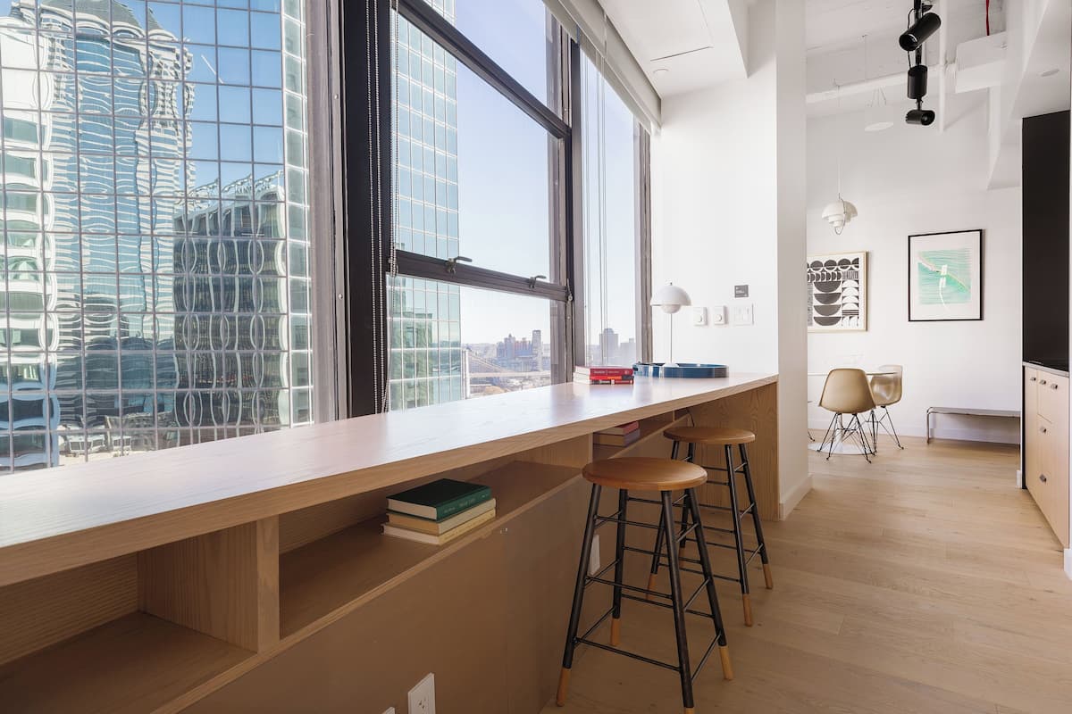 A modern kitchen with a large window, wooden bar stools, and a view of tall buildings.