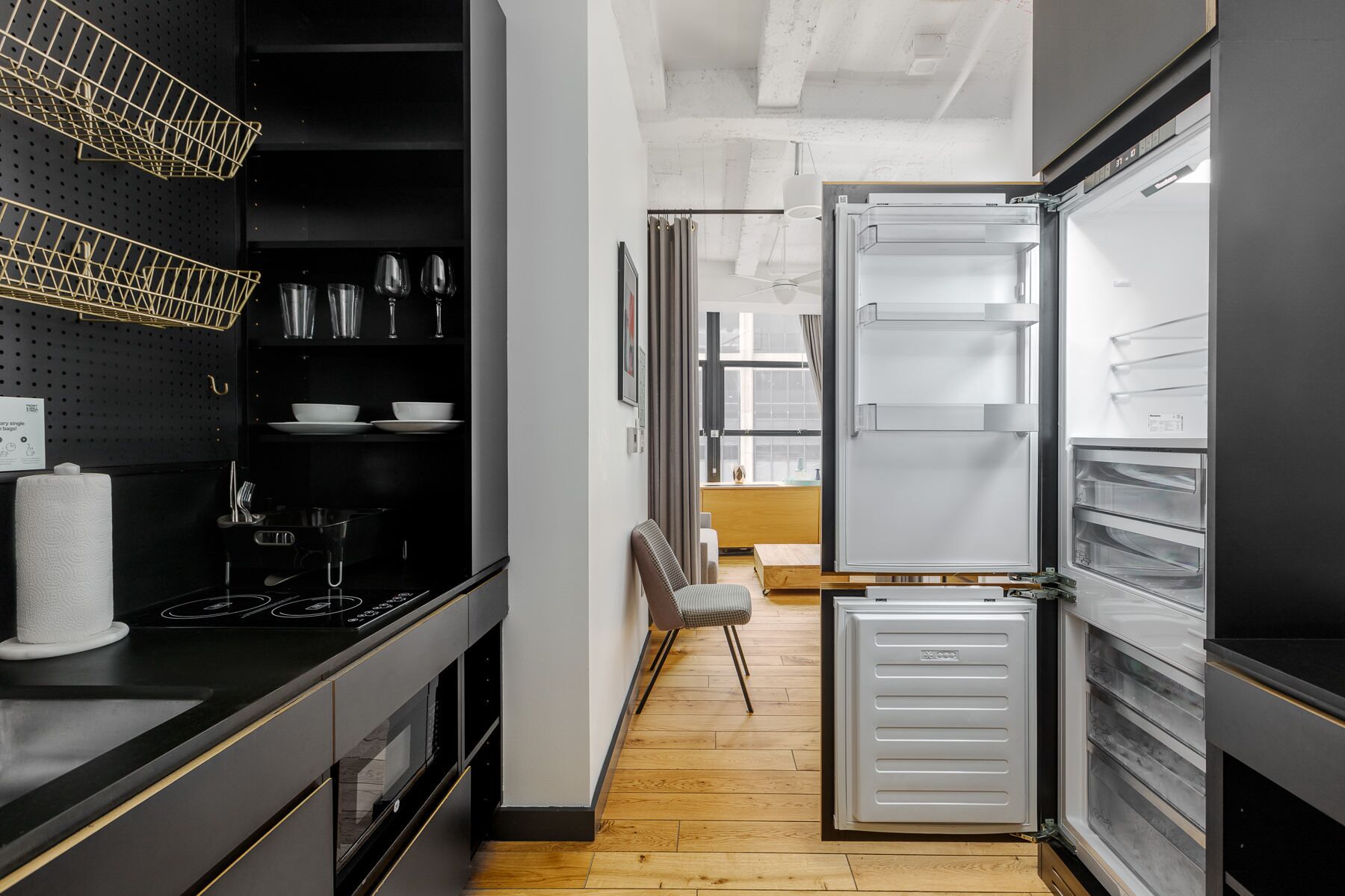 A modern kitchen with black cabinetry, a stainless steel range hood, and a built-in oven. There is a wooden dining table with a chair, and a white refrigerator with multiple shelves.