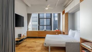 A modern hotel room with a wooden floor, a bed with a grey upholstered bench, a TV mounted on the wall, and a window with curtains.