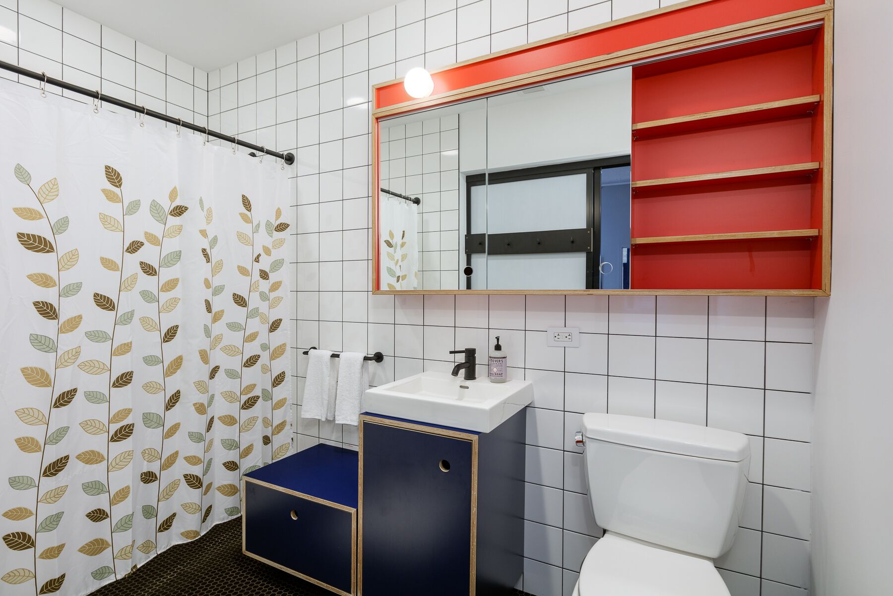 Un baño con pared de azulejos blancos, un inodoro blanco, un mueble azul, un lavabo blanco, un espejo y una cortina de ducha con estampado de hojas.