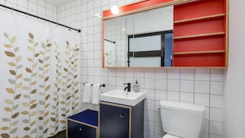 Une salle de bain avec un mur recouvert de carreaux blancs, des toilettes blanches, un meuble bleu, un lavabo blanc, un miroir et un rideau de douche orné d’un motif de feuilles.