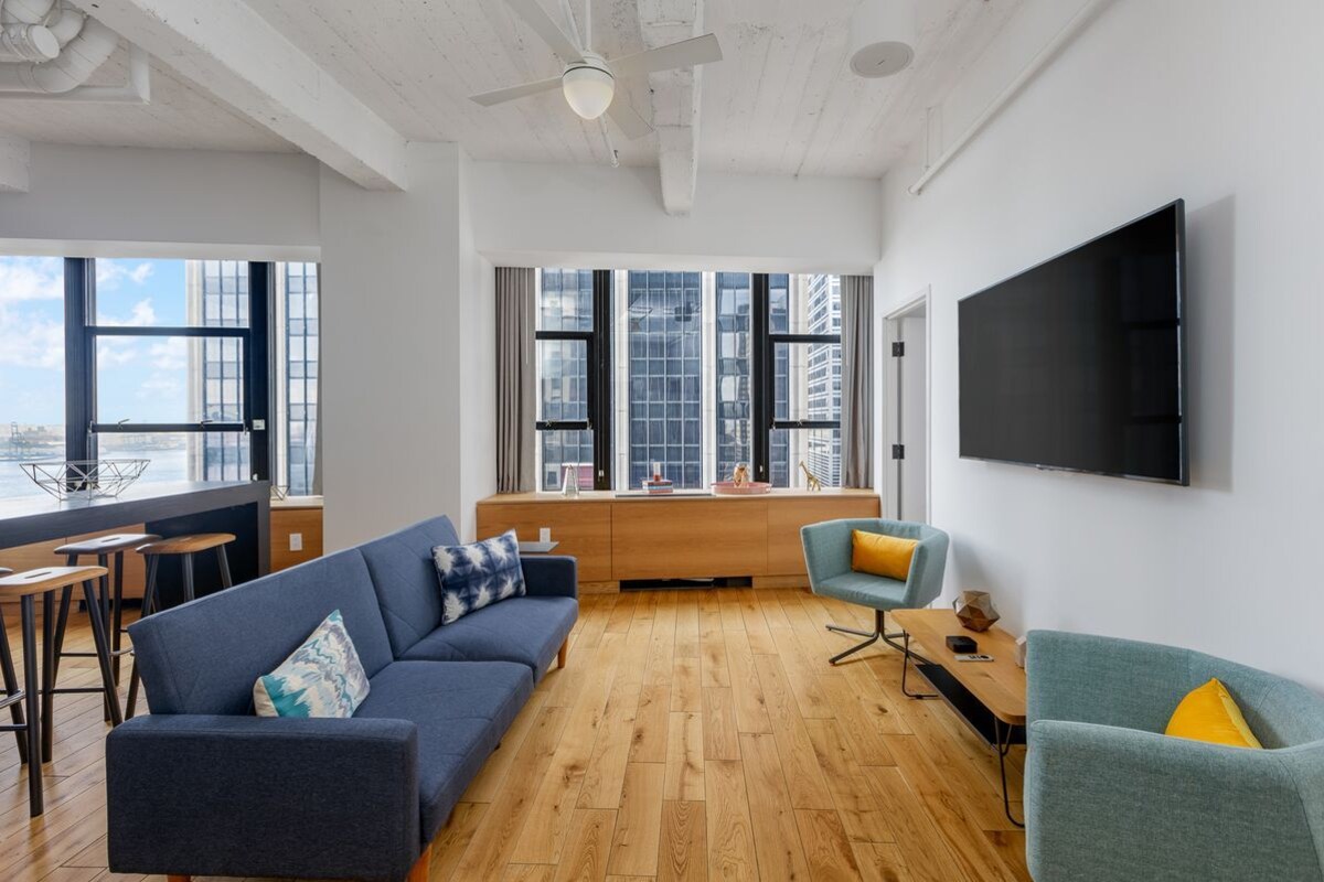 A modern living room with a sofa, armchair, and dining area, large windows, and a flat-screen TV.