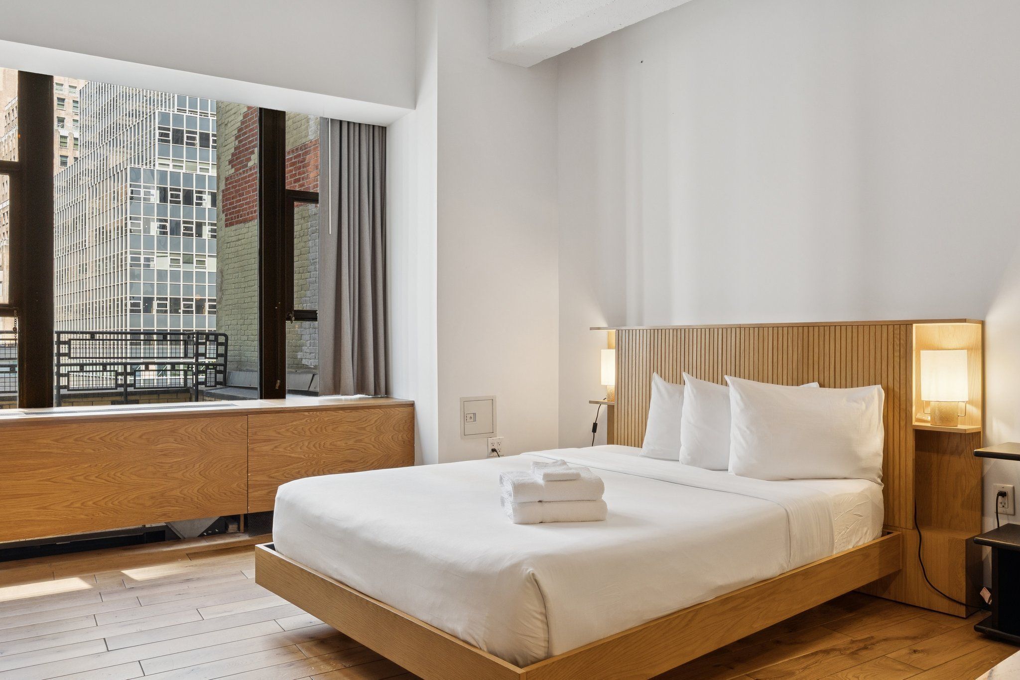 A modern hotel room with a large bed, wooden headboard, and a view of a cityscape through a large window.