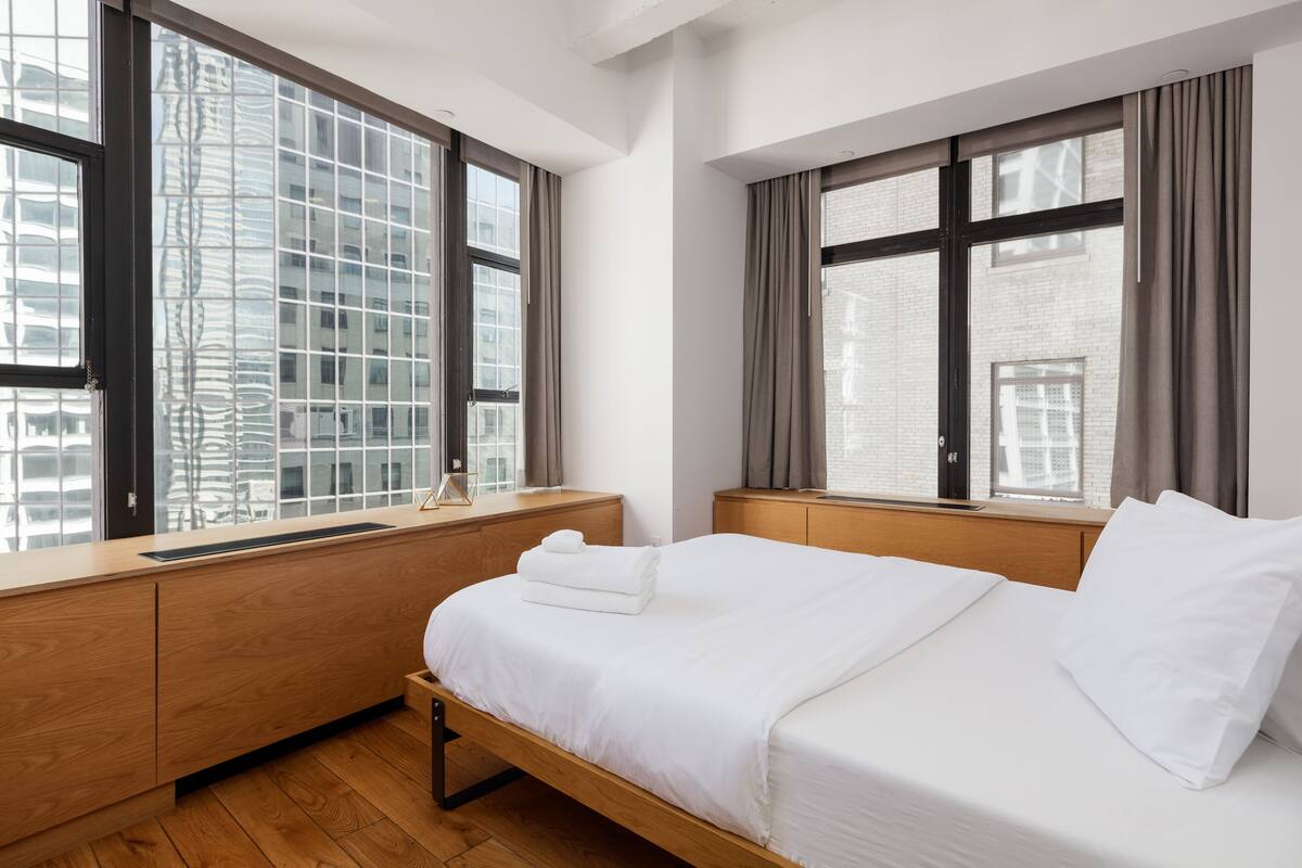 A modern hotel room with a wooden bed, a wooden nightstand, and large windows offering views of a cityscape.