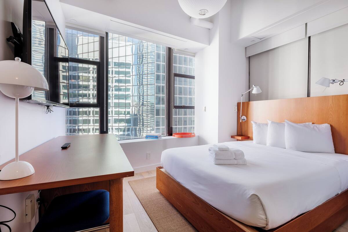 A modern hotel room with a large bed, a desk with a lamp, a view of the city through large windows, and a wooden headboard.